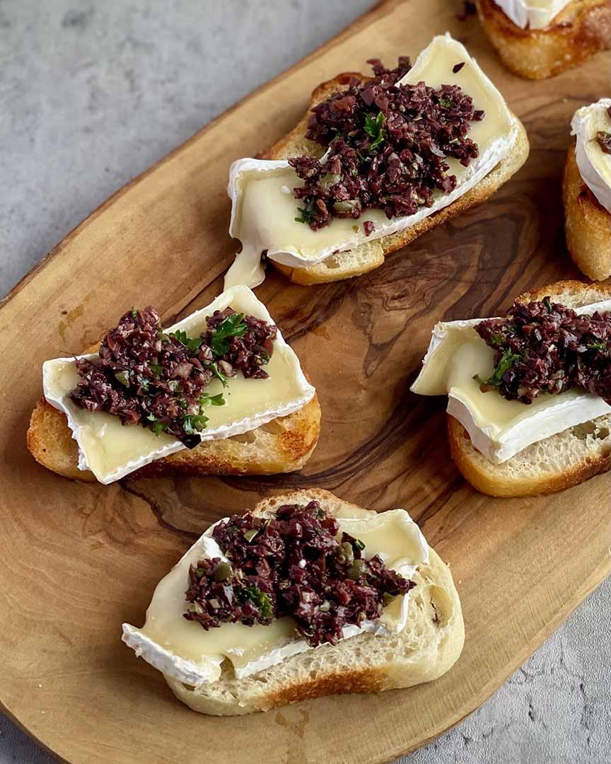 Overhead shot of olive tapenade crostini on a wooden platter outdoors
