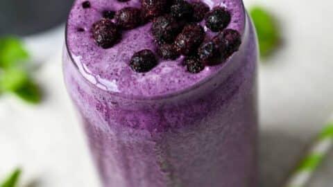 a person holding a glass of blueberry protein shake outdoors with a blurred background of mountains