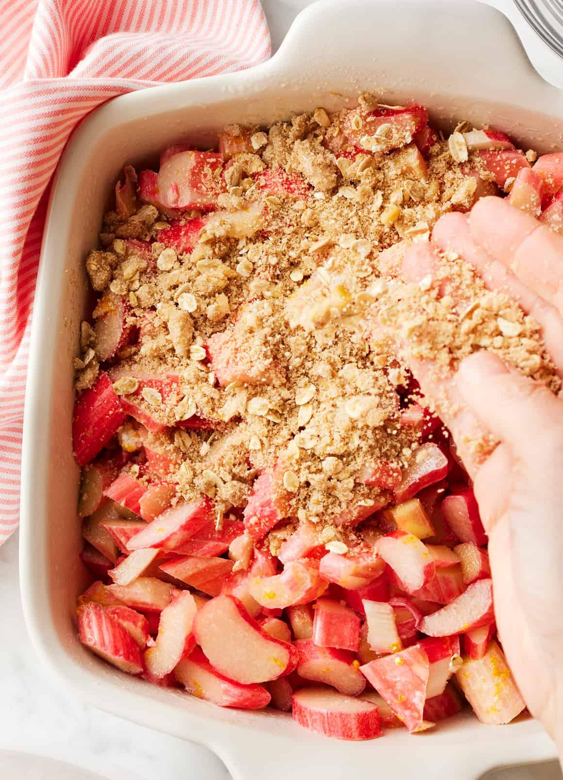a hand sprinkling crumble topping over a rhubarb filling in a baking dish