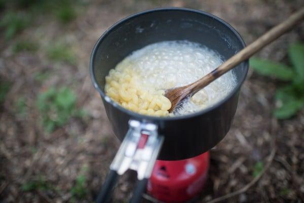 A person stirring Backcountry Mac and Cheese with Bacon over a campfire.