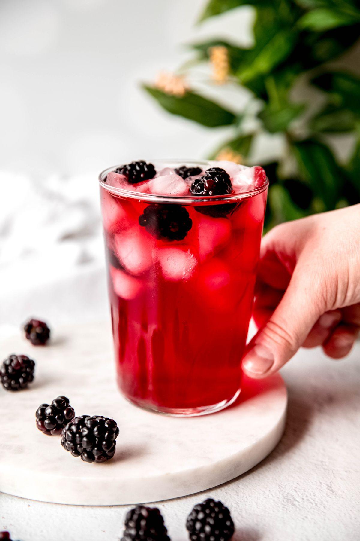 A glass of Matcha Blackberry Sage Refresh with ice, blackberries, and sage sprig garnish, on a rustic wooden table with a blurred outdoor background.