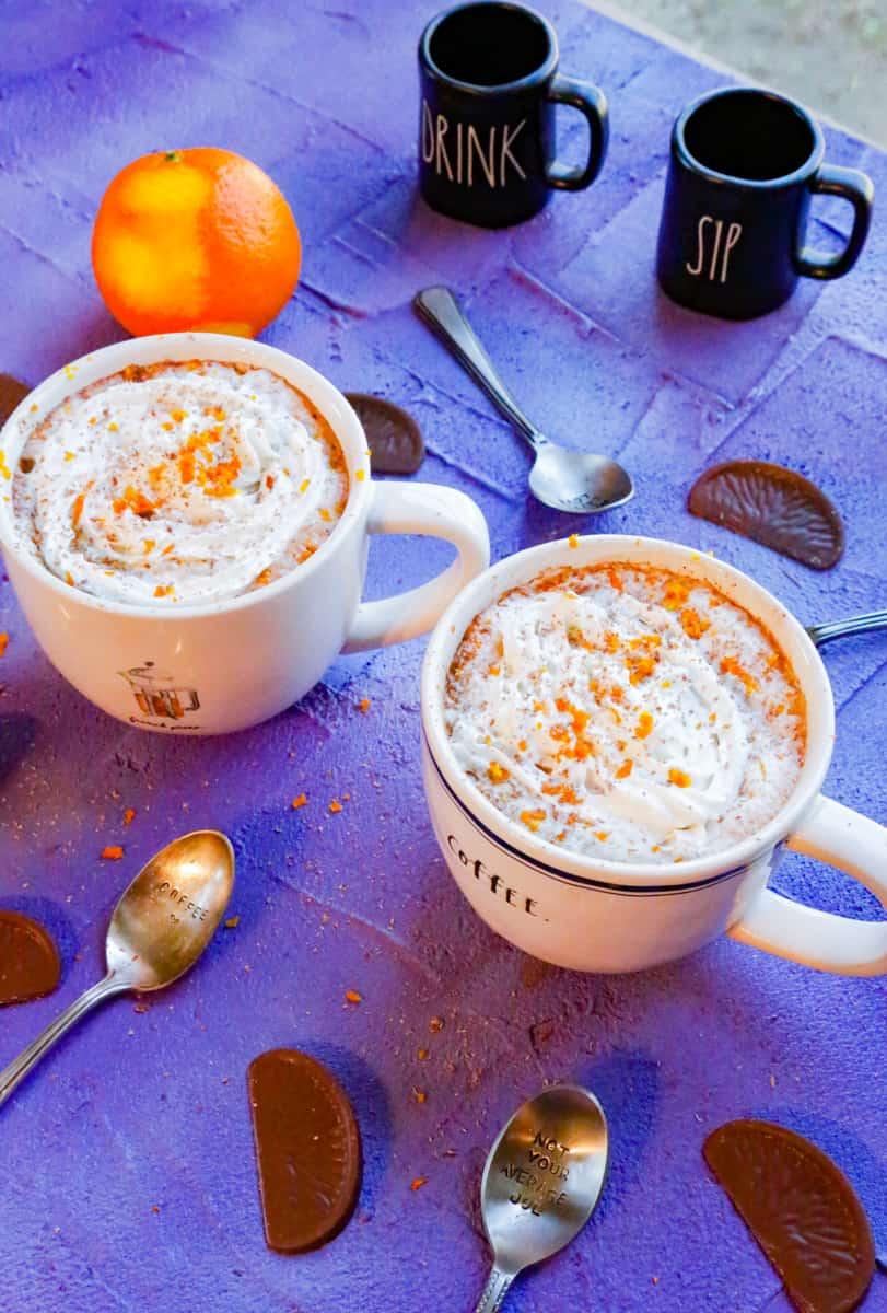 Overhead shot of two mugs of orange hot chocolate, one with whipped cream and one with marshmallows, on a rustic wooden table with orange slices