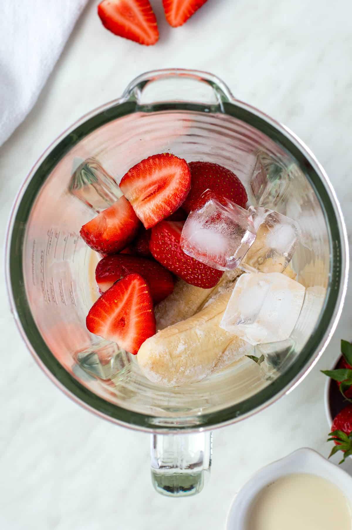 A person adding strawberries to a blender filled with banana slices and milk.