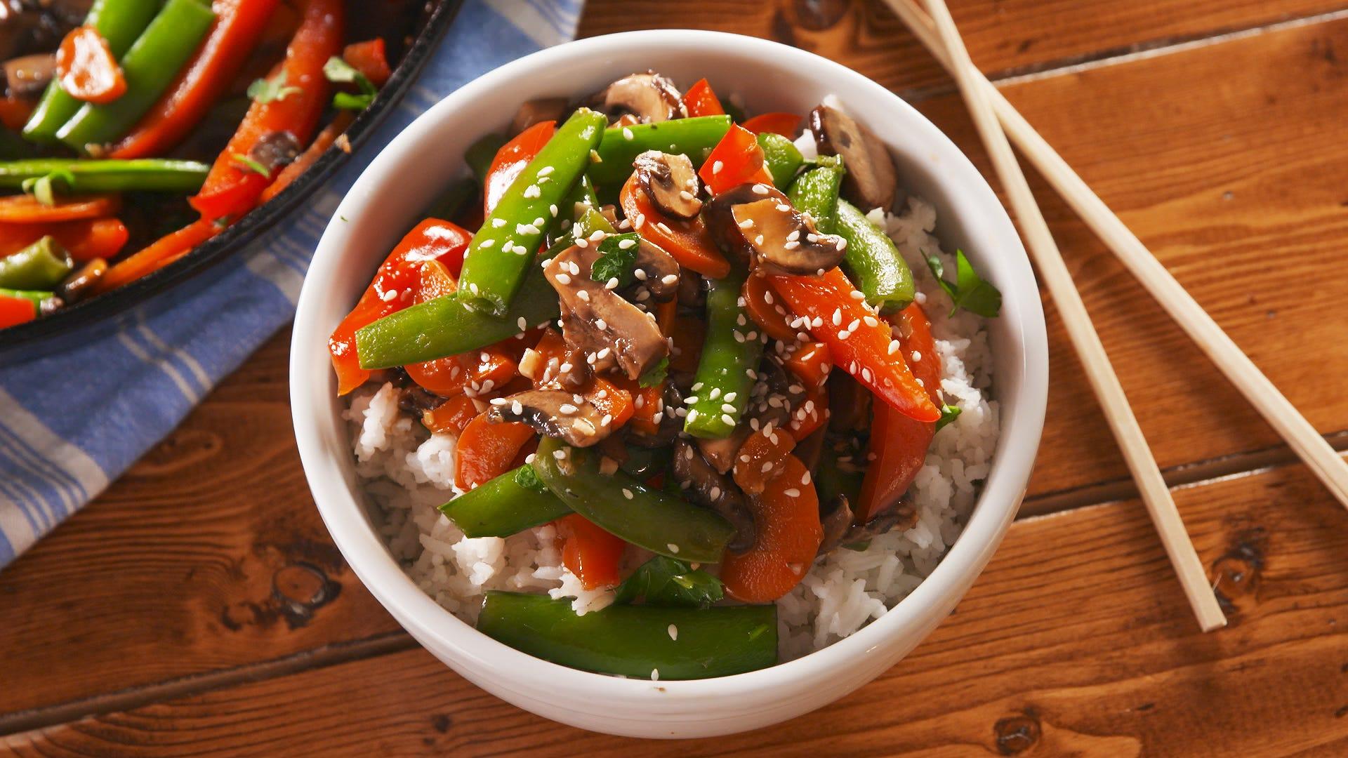 A vibrant vegetable stir-fry with visible pieces of crystallized ginger and sesame seeds, served in a white bowl.