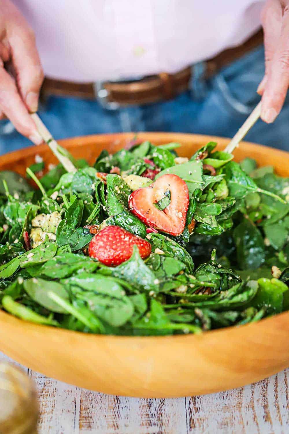 Overhead view of a person tossing a strawberry spinach salad in a large bowl.
