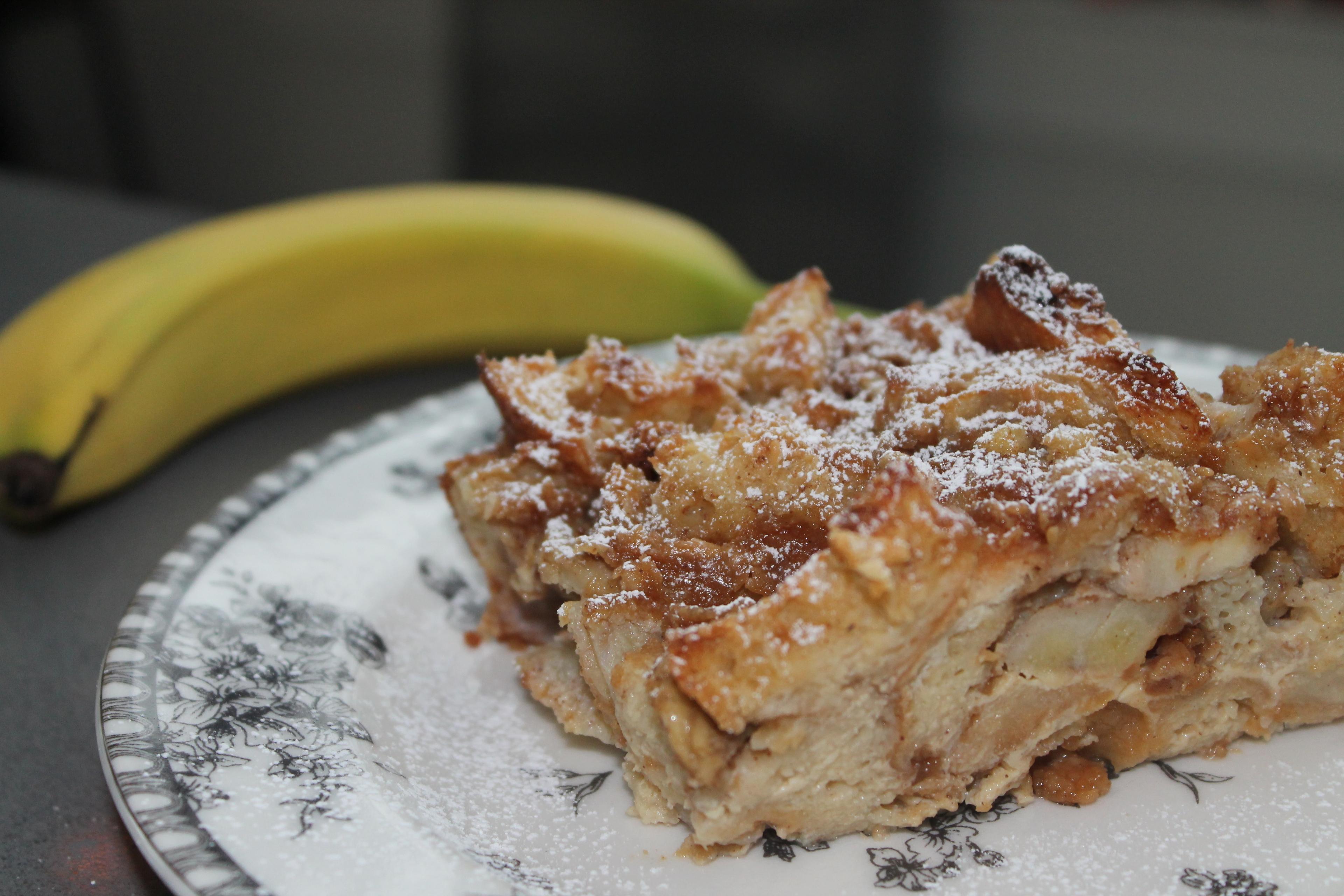 Ingredients for peanut butter banana bread pudding laid out on a wooden countertop, including bread cubes, bananas, eggs, milk, peanut butter, and spices