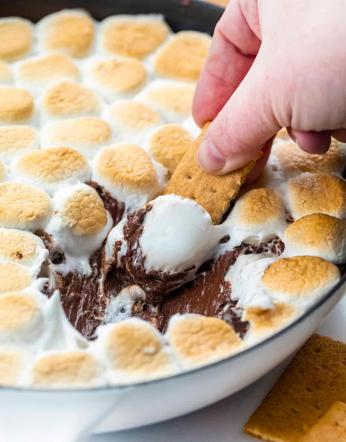 close-up shot of a graham cracker being dipped into the s'mores dip, with melted chocolate and toasted marshmallows visible