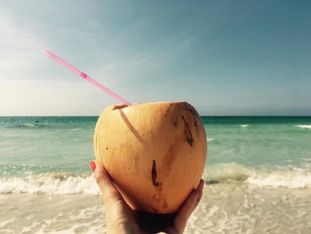 A close-up of a hand holding a Coconut Lime Hugo cocktail against a sunset beach backdrop
