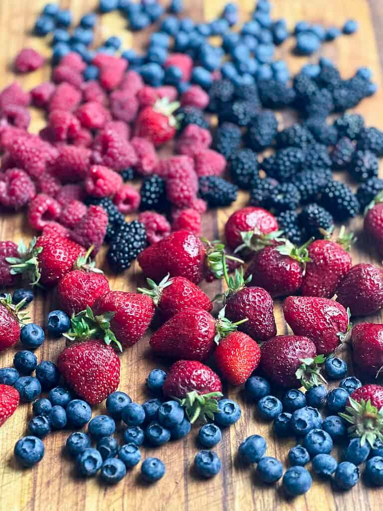 a variety of fresh summer berries (strawberries, blueberries, raspberries, blackberries) arranged beautifully on a wooden cutting board