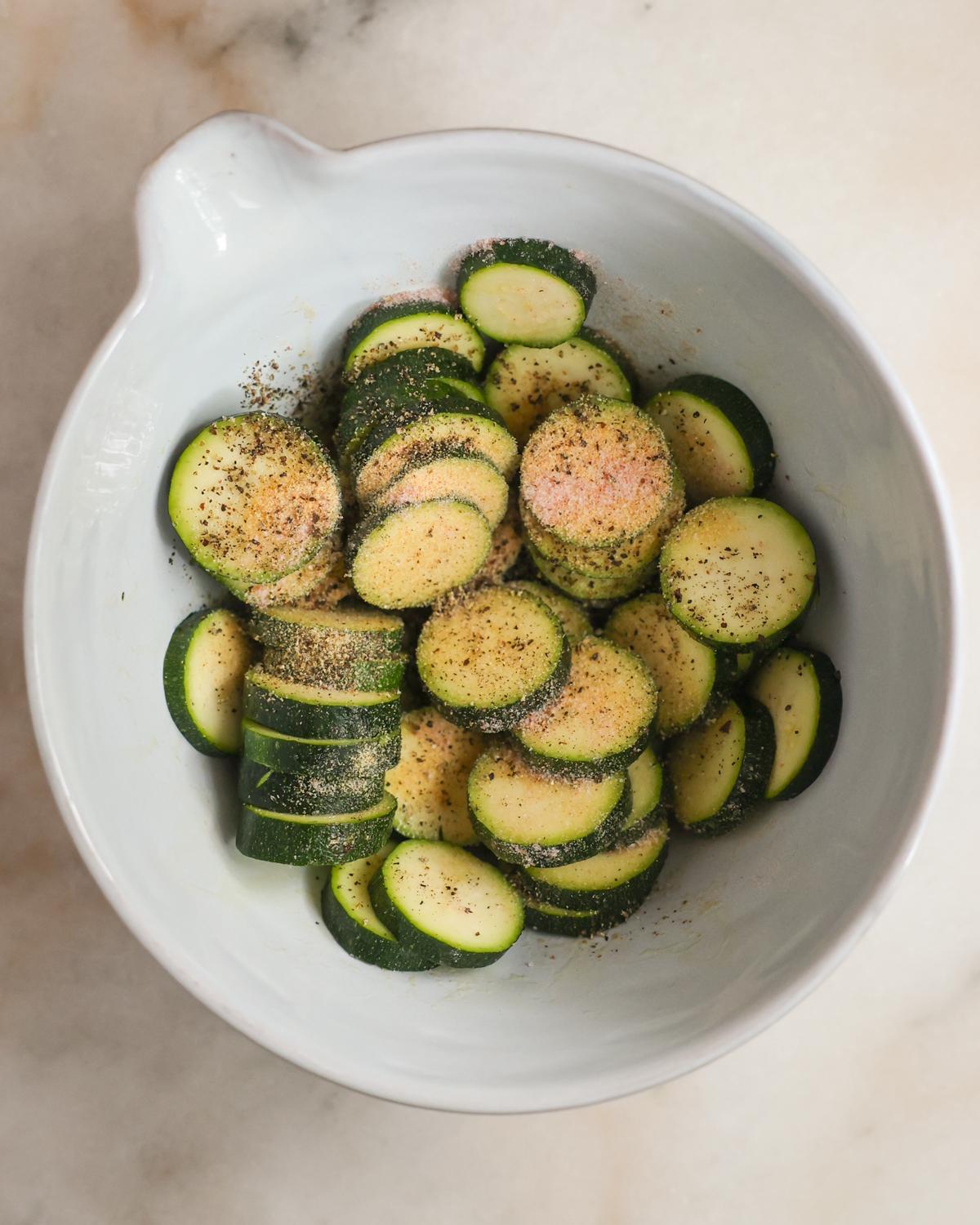 sliced zucchini being tossed with spices in a bowl
