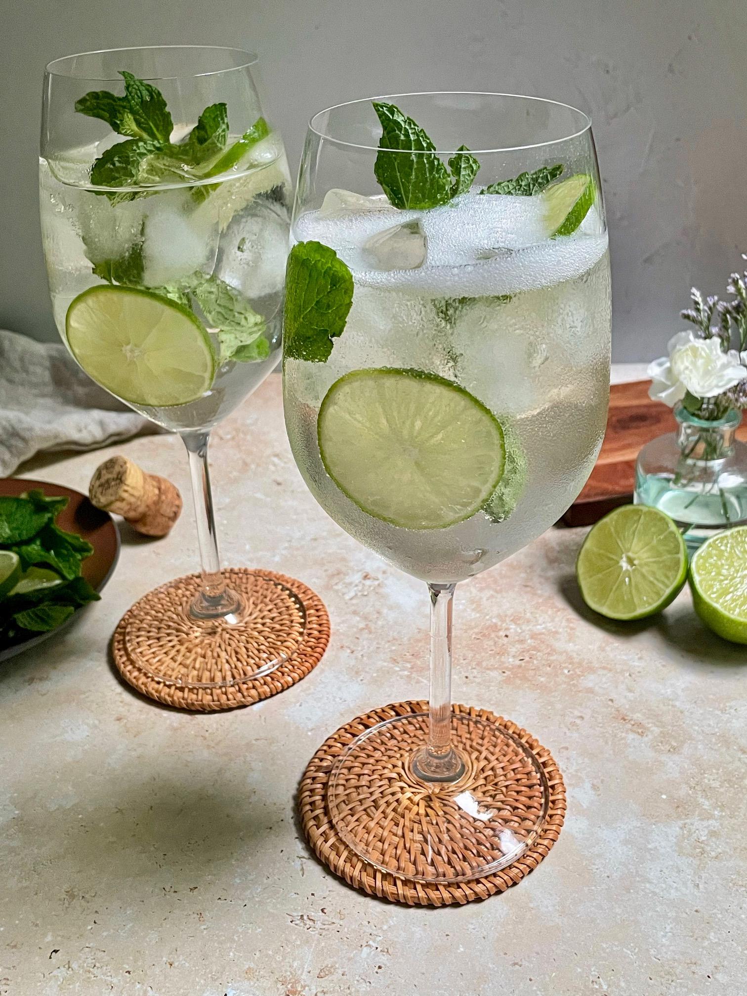 A refreshing Coconut Lime Hugo cocktail in a highball glass, garnished with lime wedges and mint sprigs, with a tropical beach background