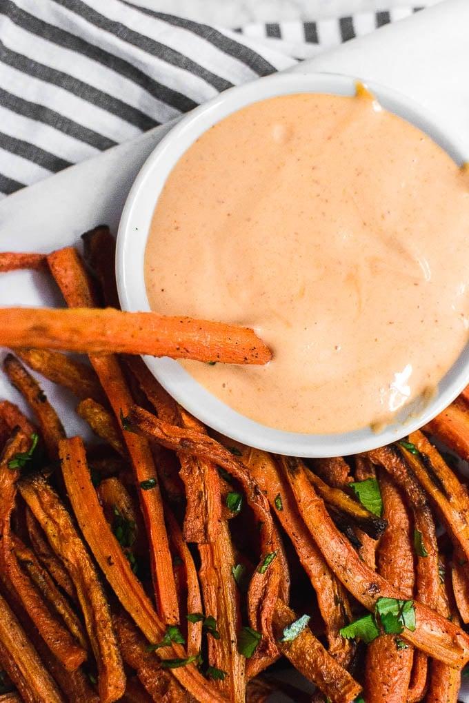 Close-up of a hand dipping a carrot fry into a bowl of vibrant sriracha mayo.