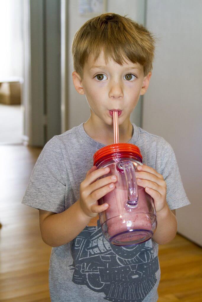 a child smiling while drinking a strawberry banana smoothie with a straw