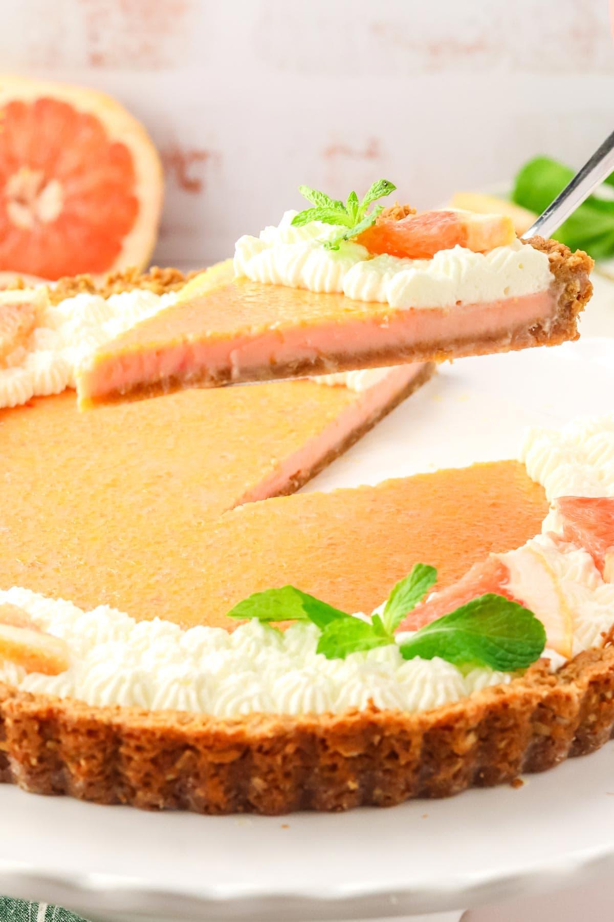 A slice of the Grapefruit & Thyme Tart being lifted from the whole tart, showcasing the flaky crust and juicy grapefruit filling
