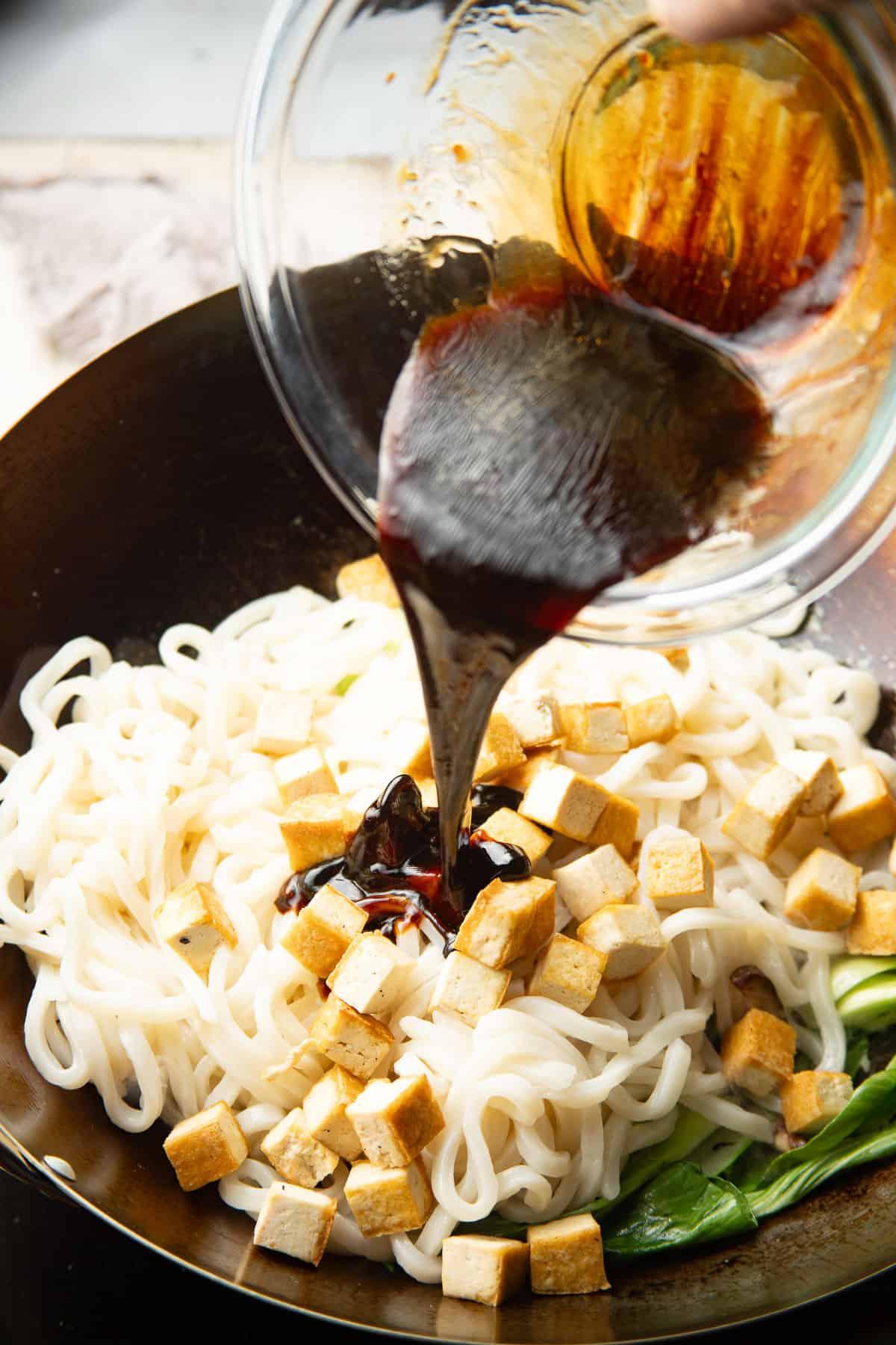 pouring sauce over tofu and udon noodles in a wok