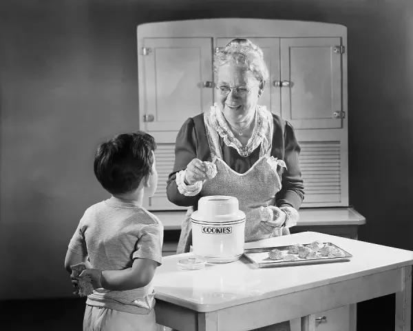 Child watching grandmother paint butter cookies, warm kitchen with baked goods