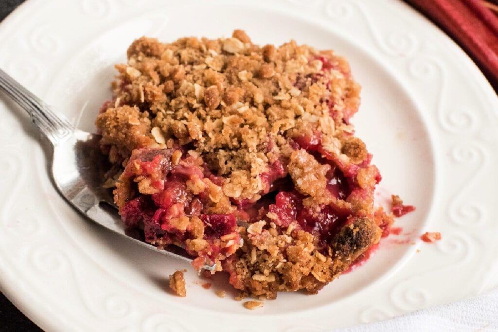 close-up shot of a slice of rhubarb crumble on a plate with a fork, showing the layers of crust, rhubarb filling, and crumble topping