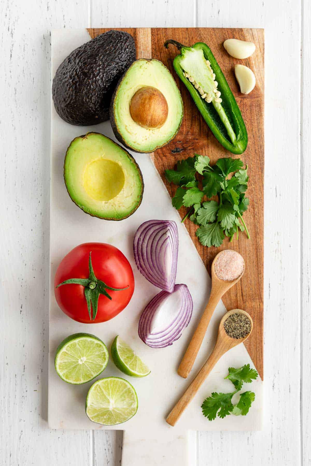 ingredients for guacamole displayed on a wooden cutting board, including avocados, lime, cilantro, red onion, and jalapeno