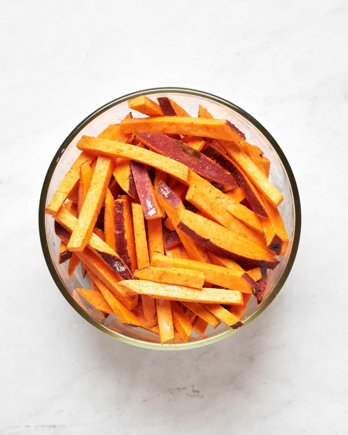 sweet potato fries being tossed in a bowl with olive oil and spices