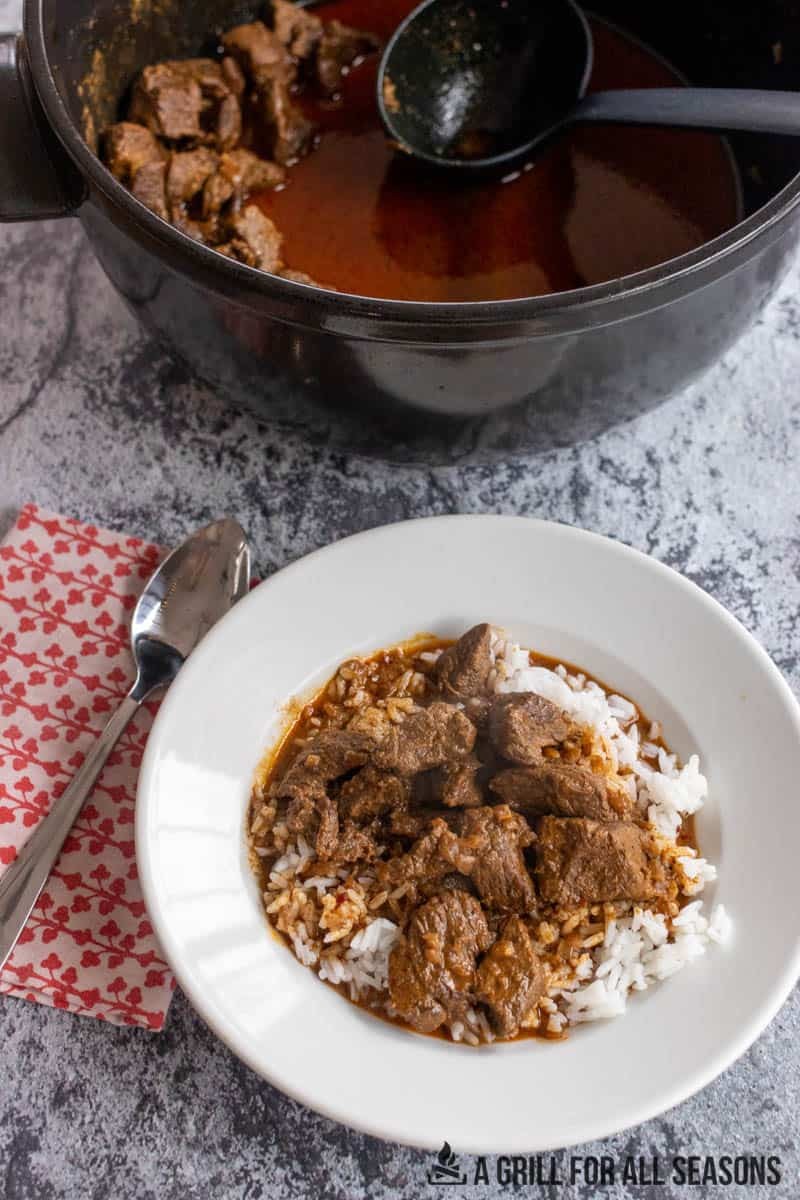 A close-up shot of a steaming bowl of Chili-Masala Beef Stew, garnished with fresh cilantro, on a rustic wooden table with a side of basmati rice and naan.