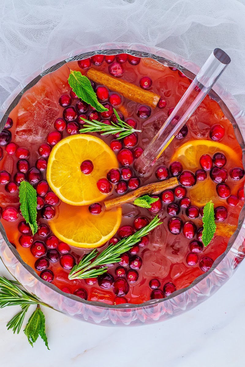 vibrant holiday spiced rum punch with orange slices, star anise, cinnamon sticks, and cranberry garnish in a large glass punch bowl, surrounded by festive twinkling lights and pinecones