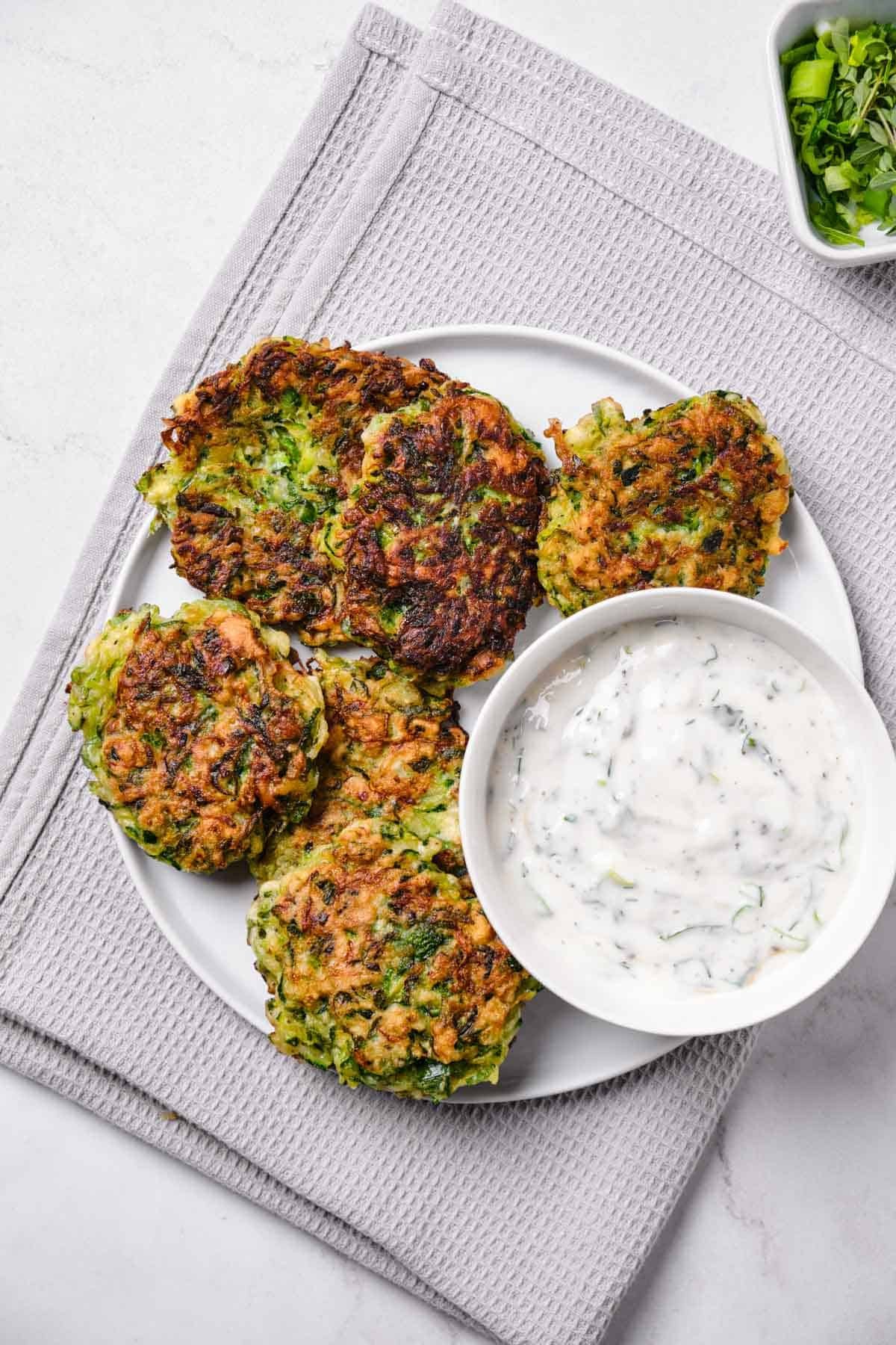 Crispy baked zucchini fritters served with a bowl of green herb yogurt dip, fresh dill, mint, and lemon wedges, bright natural light