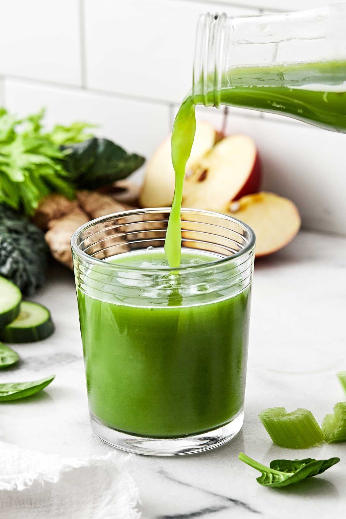 bright green juice being poured into a glass, with fresh apples, ginger, and greens surrounding it