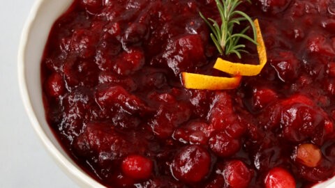 close-up photo of ruby red homemade cranberry sauce with a vanilla bean pod, in a rustic serving bowl, garnished with fresh cranberries and rosemary sprigs