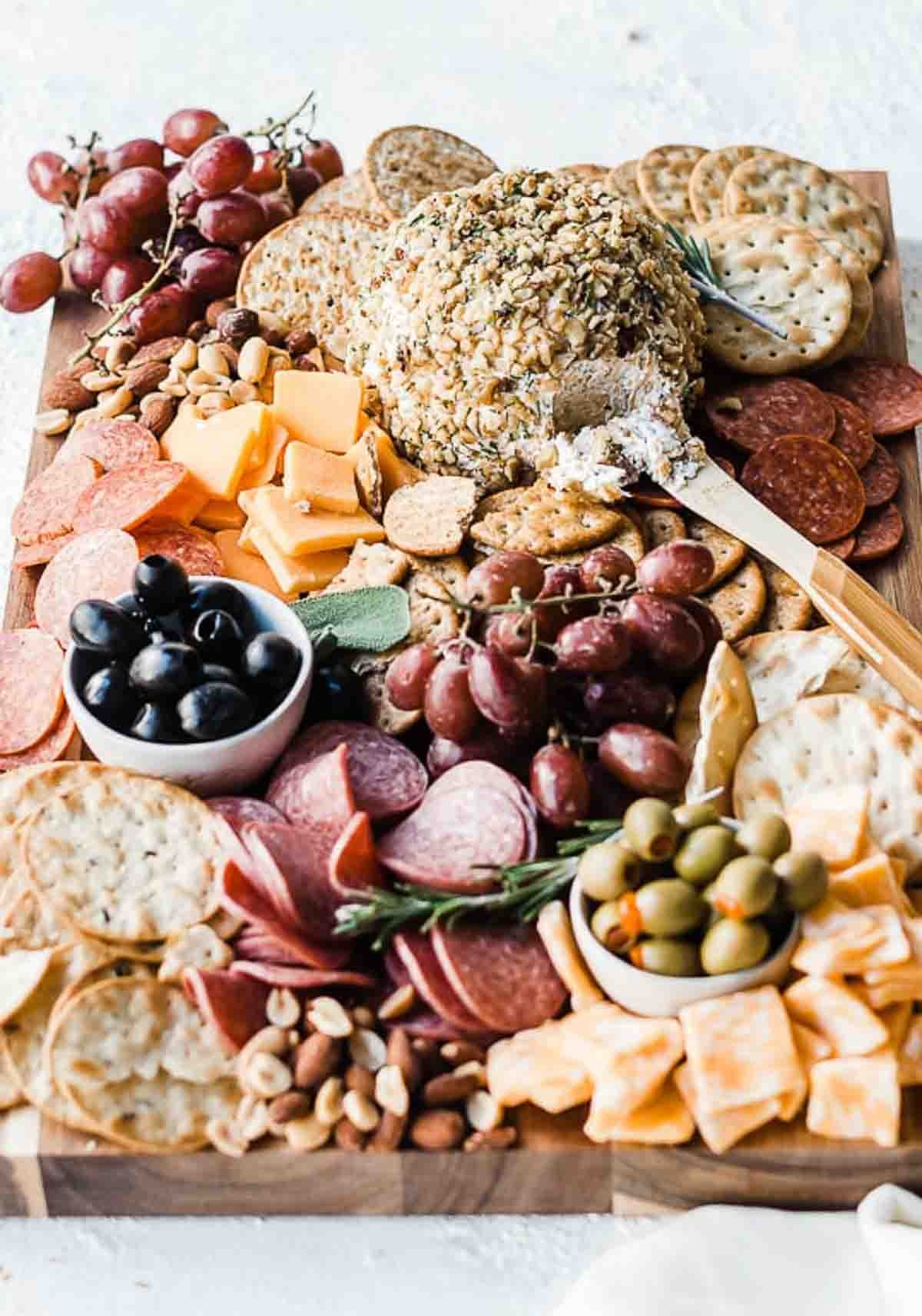 A beautifully presented herbed cheese ball coated in mixed nuts, surrounded by crackers and grapes on a rustic wooden board.