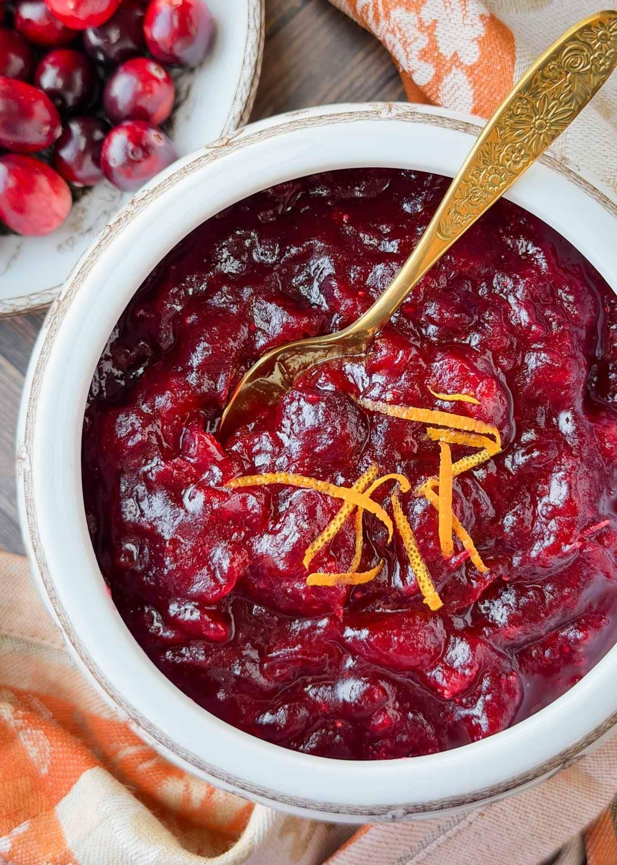 Vibrant homemade cranberry sauce in a rustic bowl, garnished with fresh cranberries and orange zest, a wooden spoon resting nearby, warm lighting