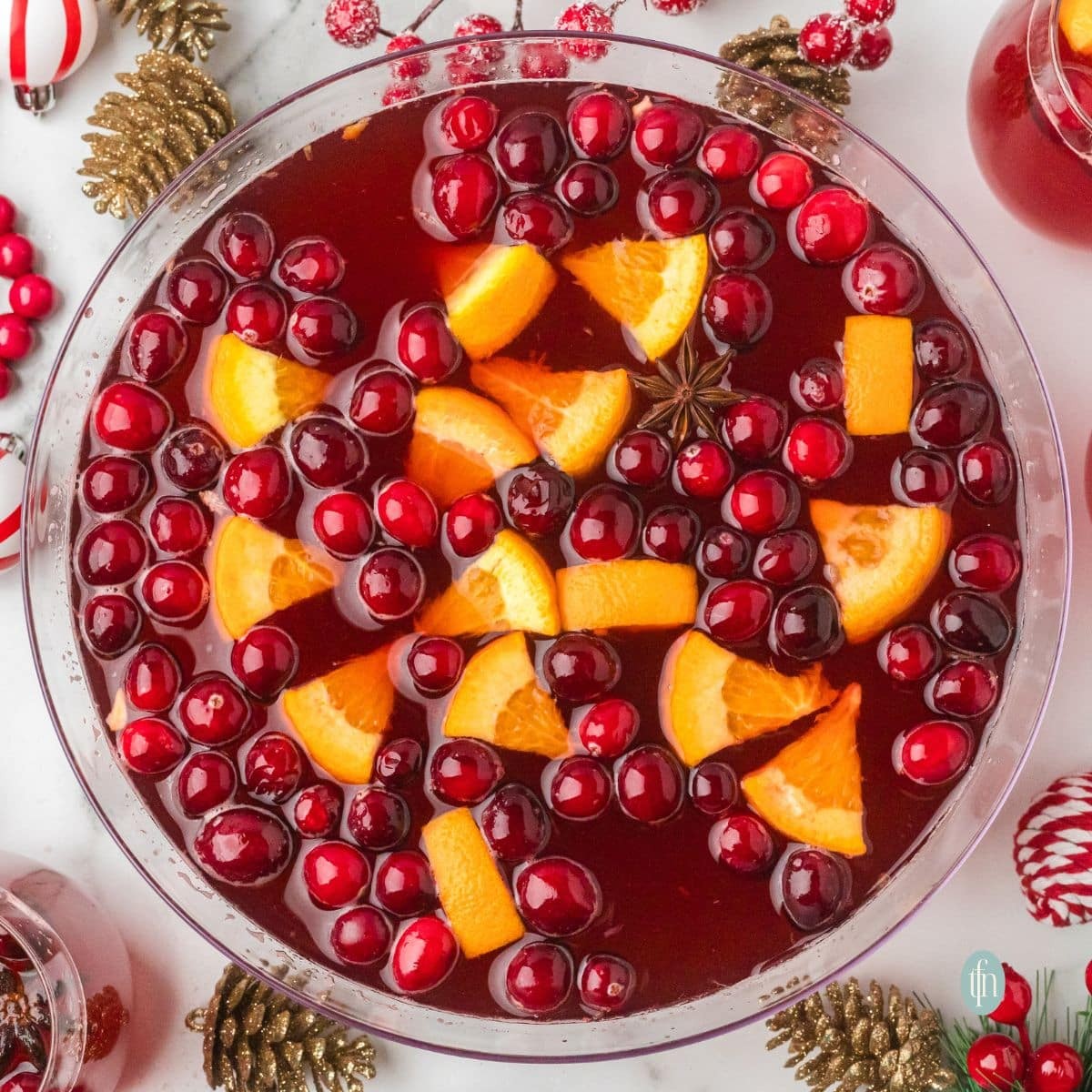 Festive orange clove punch in a large glass dispenser with holiday decor, garnished with fresh cranberries and orange slices