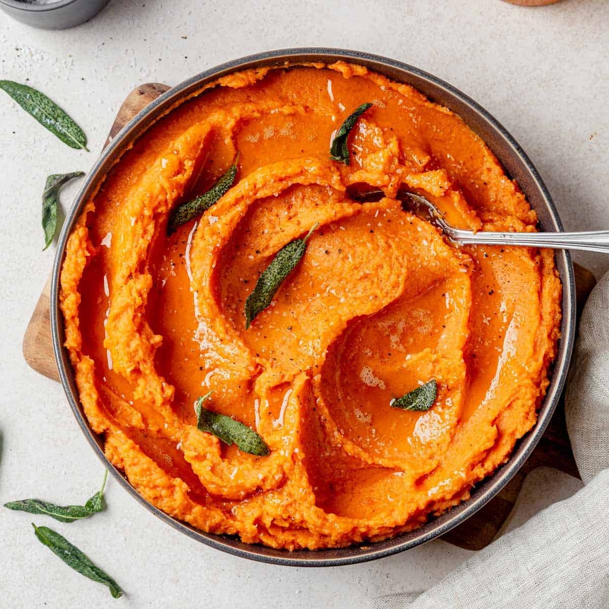 A close-up shot of a rustic ceramic bowl filled with golden brown butter sweet potato mash, garnished with fresh herbs and a drizzle of melted brown butter, on a wooden table with autumn leaves.
