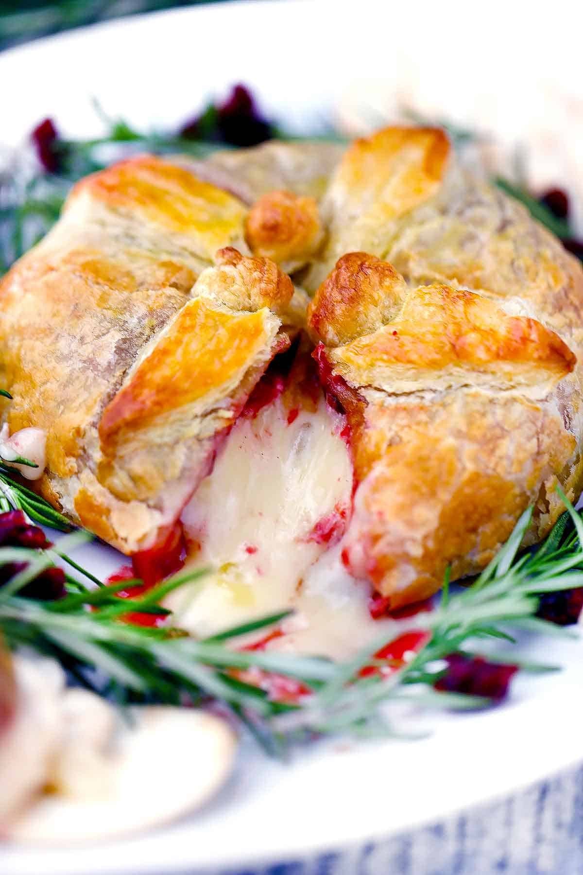 Close-up of golden brown baked brie in puff pastry with cranberry sauce and rosemary garnish, holiday setting