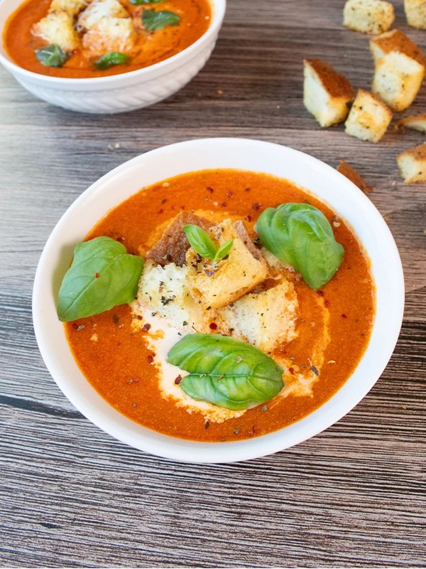 close-up shot of creamy roasted red pepper and tomato soup in a elegant white bowl, garnished with fresh basil leaves, next to rustic bread