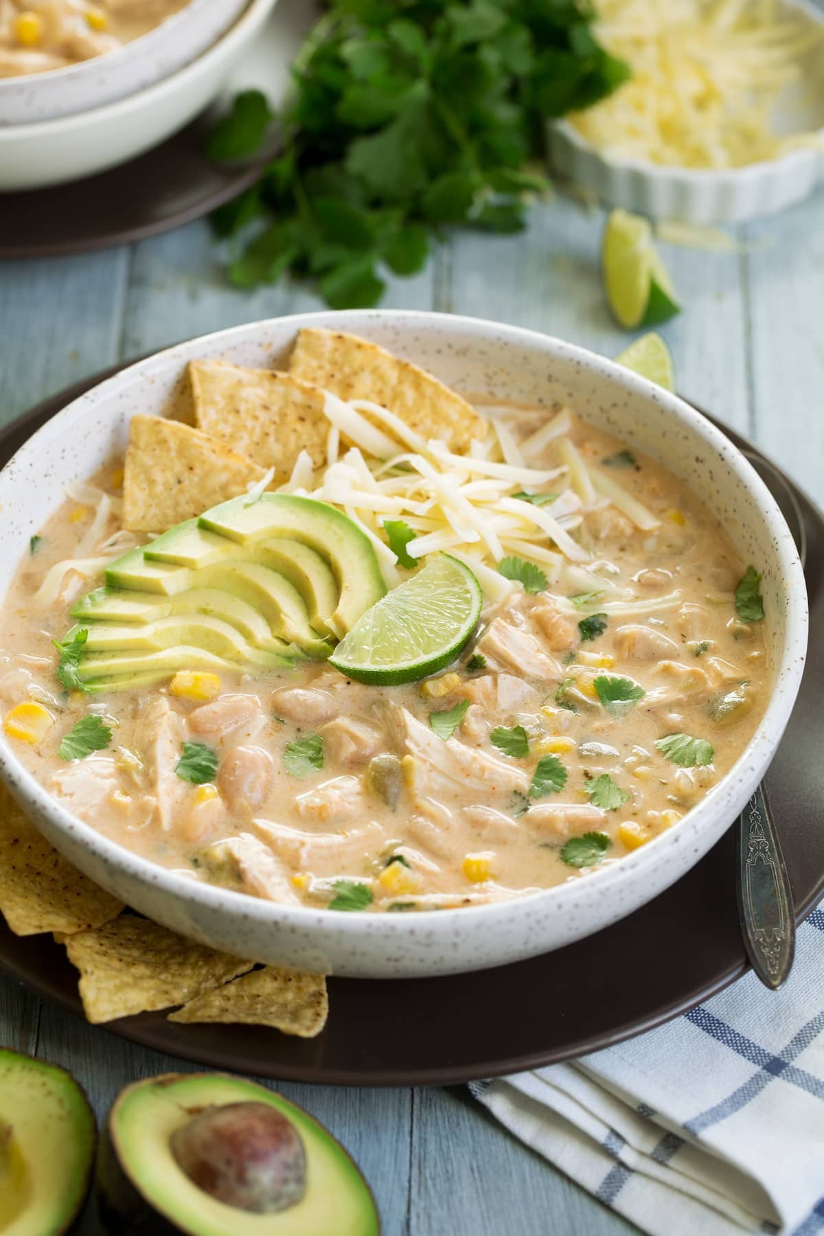 A steaming bowl of white chicken chili, garnished with fresh cilantro, a dollop of sour cream, and roasted corn kernels visible, on a rustic wooden table with a cozy blanket in the background, warm light.