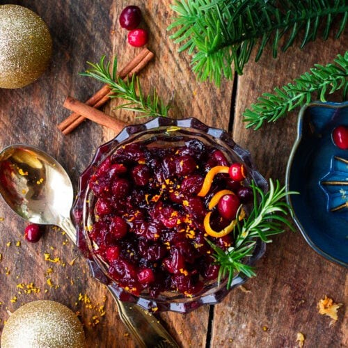 Vibrant homemade cranberry sauce in a decorative bowl, fresh cranberries and orange zest in background, festive table setting.