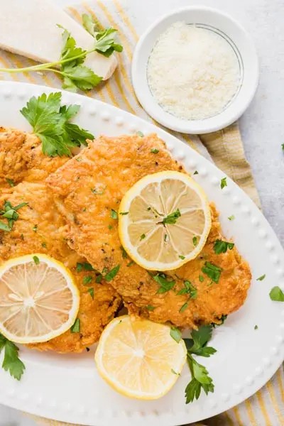 golden crispy parmesan chicken cutlets on a plate with lemon wedges and parsley