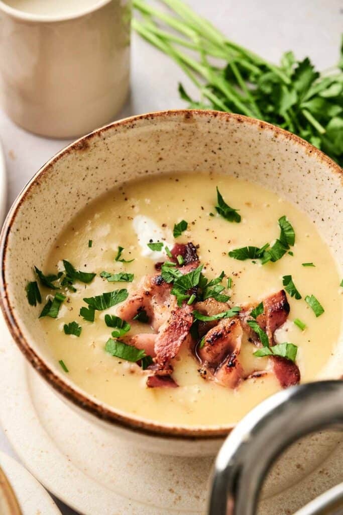 Creamy potato soup in a rustic bowl, topped generously with crispy bacon bits and fresh green scallions, cozy setting